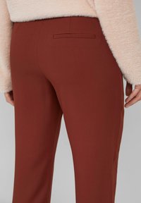 Persoon in hooggewaisted roestkleurige pantalon met een enkele achterzak, gecombineerd met een lichtroze fuzzy trui.