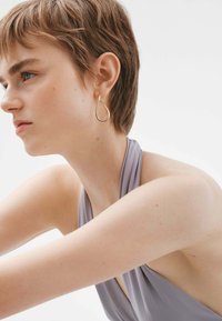 Pendiente en forma de lágrima de oro colgando de la oreja. Textura suave y lisa. Top halter gris con tela drapeada. Fondo neutro.