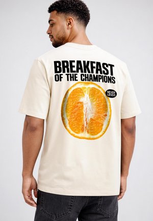 Mann trägt beige T-Shirt mit großer Orangenscheibe und dem Schriftzug „BREAKFAST OF THE CHAMPIONS“ auf dem Rücken.
