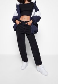 Person klädd i en marinblå och vit pufferjacka, svart crop top, lösa svarta jeans och vita sneakers, stående med händerna i fickorna.