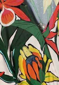 Tissu floral lumineux avec de grands motifs de fleurs et feuilles tropicales rouges, jaunes, orange et vertes sur un fond clair.