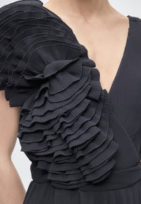 Μαύρο φόρεμα με βαθύ V λαιμόκοψη και μεγάλο στρωματοποιημένο πιέτα στολισμένο βολάν σε έναν ώμο.