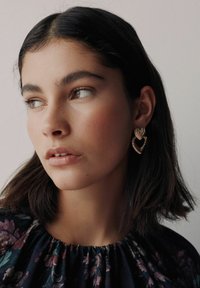 Gold-tone earrings featuring a heart shape with a textured, ribbed design. The model has shoulder-length dark hair and wears a floral top.
