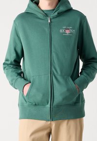 Felpa verde con zip realizzata in materiale morbido, con logo frontale, tasche laterali e polsini a coste. Abbinata a pantaloni beige.