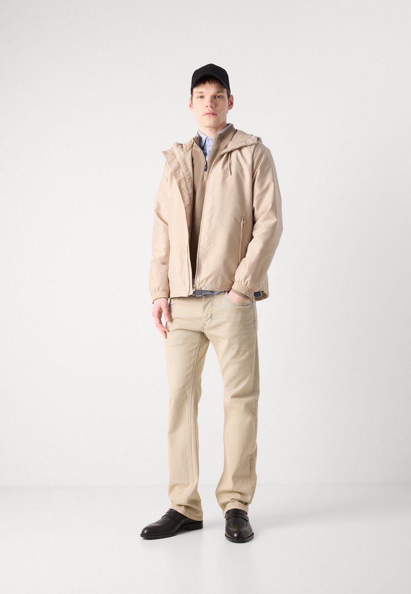 Veste à capuche beige avec des poignets élastiques sur un pull en tricot beige, portée avec un jean beige clair et des chaussures en cuir noires. Casquette noire unie.