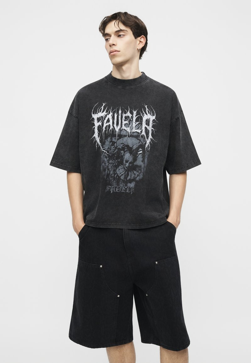 Junger Mann trägt ein gewaschenes schwarzes oversized Grafik-T-Shirt und schwarze weite Shorts, steht mit den Händen in den Taschen und schaut nach links.