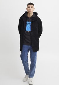 Sort vandafvisende jakke med hætte, parret med en mørkegrå hoodie med en blå grafisk illustration og tekst, samt lyseblå jeans.