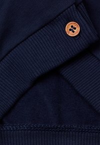 Námorně modrý svetr s vroubkovanými manžetami a lemem, který má měděný knoflík se čtyřmi otvory, hladkou texturu a detaily šití.