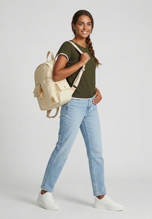 Nuori nainen, jolla pitkä letti, vihreä t-paita, vaaleat farkut, valkoiset lenkkarit, kantaa beigeä reppua yhdellä olkapäällä ja hymyilee.