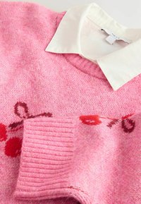 Rosa Strickpullover mit rundem Ausschnitt, darunter ein weißes Kragenhemd. Der Pullover hat eine weiche Textur und florale Muster in Rot und Burgunder.