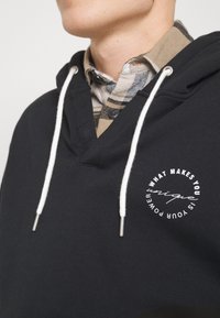 Felpa nera con cappuccio e lacci bianchi, presenta un scollo a V e una grafica circolare bianca sul davanti, indossata sopra una camicia a quadri.