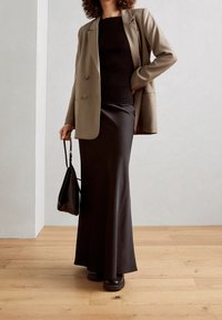 Frau trägt beigen Blazer über schwarzem geripptem Top und langem schwarzen Rock, hält schwarze Handtasche und steht auf einem Holzboden vor einer schlichten Wand.