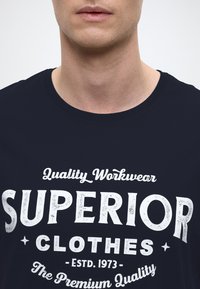 Navy blue cotton t-shirt featuring white text: "Quality Workwear SUPERIOR CLOTHES ESTD. 1973 The Premium Quality," with decorative accents.