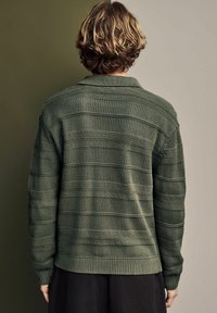 Maglione lavorato a maglia verde oliva con strisce testurizzate e orlo a coste. Il colletto ha un design rilassato. La texture del tessuto è intrecciata e spessa.