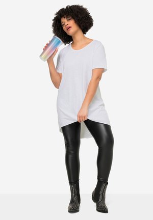 Frau mit lockigem Haar trägt ein weißes oversized Hemd, schwarze Lederleggings, schwarze Nieten-Stiefeletten und hält einen bunten, texturierten Becher.