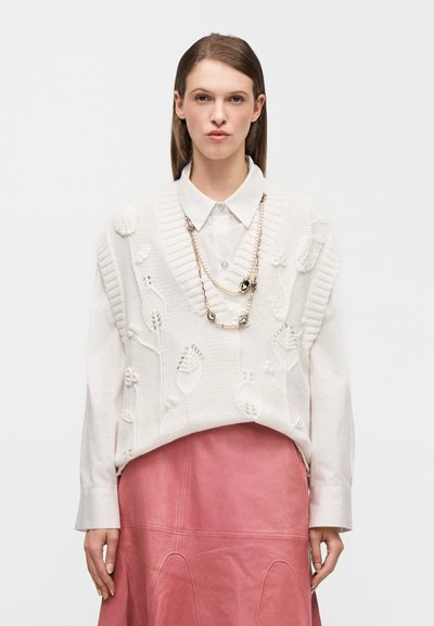 Pull blanc en maille avec motifs floraux superposé à une chemise de couleur claire, associé à une jupe en cuir rose. Collier en argent avec accents.