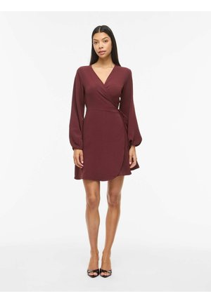 Kvinde iført en bourgognefarvet slå-om-kjole med lange ærmer og sorte åbne hæle, stående foran en ensfarvet hvid baggrund.