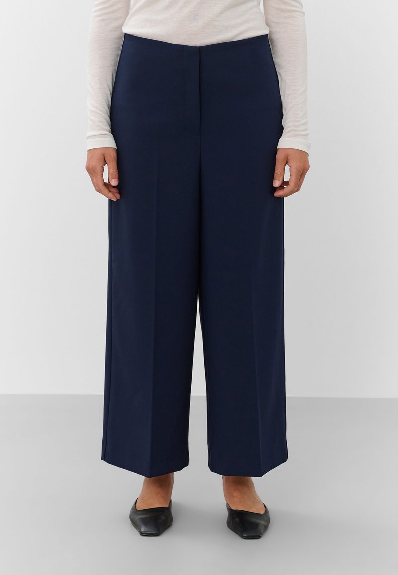 Model draagt marineblauwe wijde cropped broek met zwarte puntige platte schoenen en een licht langärmelig topje, staand tegen een effen achtergrond.
