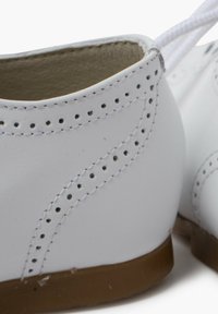 Scarpe in pelle bianca con accenti perforati, cuciture curve e suole in gomma marrone; presentano una punta arrotondata e lacci bianchi.