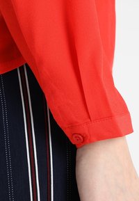 Blouse rouge légère à texture lisse, dotée d'un poignet boutonné et d'une manche retroussée. Tissu rayé bleu visible en dessous.