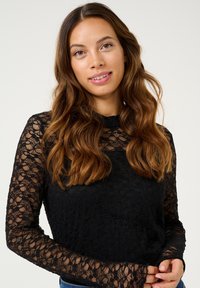 Haut en dentelle noire à manches longues avec un col montant, présentant un motif floral et une texture transparente, ajusté à la taille et aux bras.