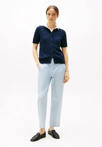 Granatowy kardigan z dzianiny o warkoczowym wzorze, z krótkimi rękawami, z zapinaniem na guziki, w zestawie z jasnoniebieskimi spodniami i czarnymi mokasynami. Gładka tekstura, luźny krój.