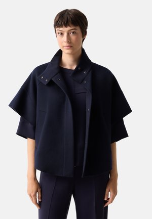 Jong persoon met kort donker haar draagt een donkerblauwe korte mouw jas over bijpassende donkere kleding tegen een effen lichte achtergrond.