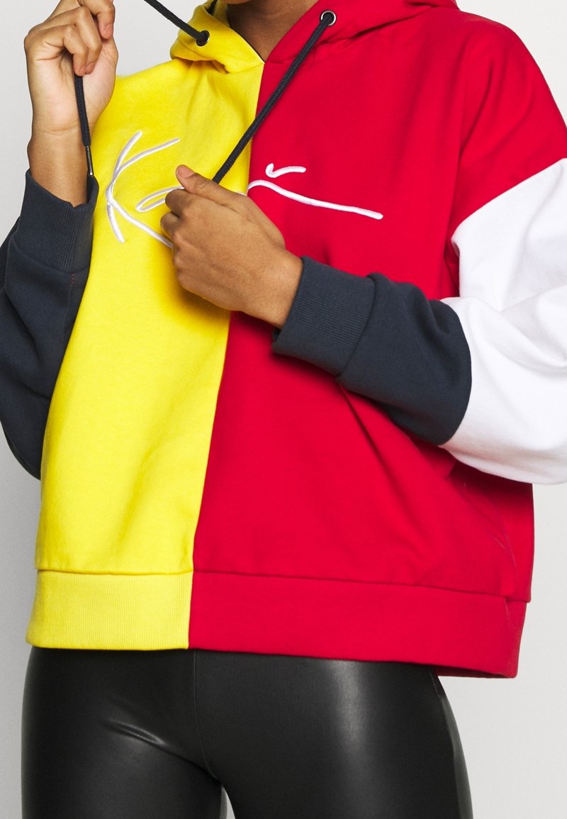 Personne portant un sweat à capuche color-block avec des sections jaune, rouge, noir et blanc, ajustant le cordon noir près du cou.
