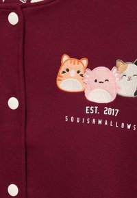 Burgundská látka s vyšitými kreslenými zvířaty: oranžově pruhovaná kočka, růžové stvoření s křídly a šedá kočka. Bílé patentky.