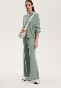 Světlově zelený pletený sportovní komplet s kapucí na zip a širokými kalhotami. Modelka drží měkkou krémovou kabelku přes rameno, obuté má bílé tenisky.