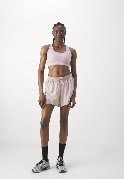 Mujer atlética de pie mirando hacia adelante, vestida con un sujetador deportivo rosa claro, pantalones cortos, calcetines negros y zapatillas blancas con detalles en azul, sobre un fondo blanco.