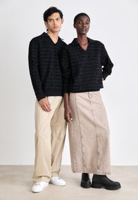 Felpe nere a trama con grande logo stampato, pantaloni beige rilassati, una lunga gonna beige e stivali platform neri. Sfondo neutro.