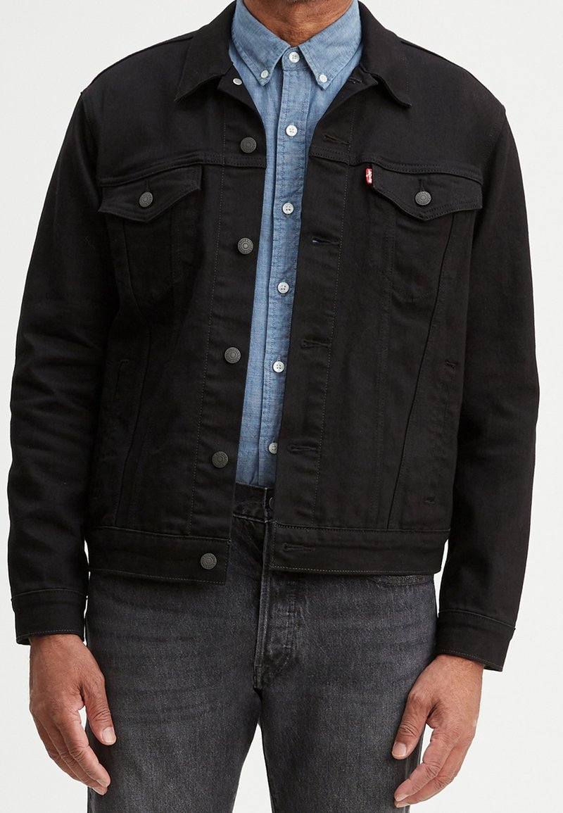 Homme portant une veste en jean noire à boutons par-dessus une chemise chambray bleue et un jean gris foncé, debout, bras détendus.