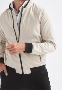 Beige Bomberjacke mit schwarzen Bündchen und Reißverschluss-Akzenten, aus glattem, leichtem Stoff mit einem schlanken Design.