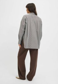 Camicia a righe grigia e bianca, dalla vestibilità ampia, con maniche lunghe, abbinata a pantaloni ampi marroni e sneaker, vista da dietro.