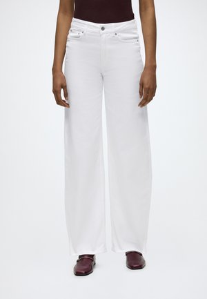 Personne portant un pantalon blanc taille haute à jambes larges, un haut foncé et des chaussures bordeaux foncé à bout carré avec un détail chaîne, debout devant un fond uni.