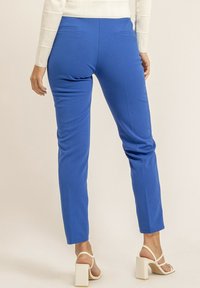 Pantalons ajustés bleus au texture lisse, coupe slim, et deux poches arrière, associés à des sandales à talons blocs blanches.