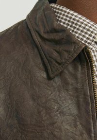 Brown leather jacket featuring a textured surface, with a folded collar and a gold zipper. Worn over a checkered fabric shirt.
