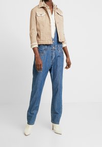 Giacca di jeans beige cropped con due tasche sul petto, chiusura a bottoni, abbinata a salopette di jeans blu dal fit morbido e con leggeri strappi.