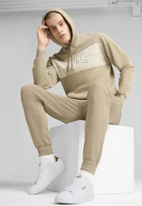 Felpa beige con logo, pantaloni jogger beige abbinati e sneakers bianche. Tessuto morbido, vestibilità rilassata e texture sottile su tutto l'outfit.