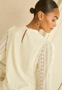 Vit spetsblus med texturerat tyg, långärmad med blommönster, nyckelhålsöppning baktill med knappdetalj och volangkantad halsringning.