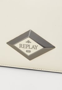 Borsa in pelle sintetica color crema con un logo metallico a forma di diamante che presenta la scritta "REPLAY" e "1981" in lettere nere.