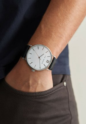 Silverklocka med rund urtavla, vit urtavla, svart läderarmband, minimalistiska timmarkeringar och en datumsdisplay vid klockan sex.