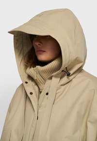 Beige vandtæt jakke med en stor hætte, lynlås foran og knapper. Bæres over en cremet ribbet turtleneck, som har en glat tekstur.