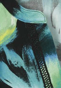 Mehrfarbige Bluse mit einer Mischung aus blauen, grünen und schwarzen farbigen Mustern, ergänzt durch filigrane schwarze Spitzenverzierungen am Rand.
