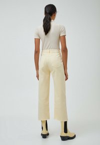 Cremig gerippter Oberteil kombiniert mit hoch taillierten, blassgelben Weitbeinhosen. Das Model trägt beige Knöchelstiefel mit schwarzen seitlichen Akzenten.