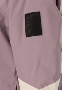 Lila ytterkläder med struktur tyg, med en svart "SLOPE STYLE" patch på ärmen och en gräddfärgad diagonalt accent.