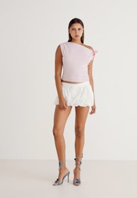 Top rosa chiaro a spalla unica con dettaglio floreale, abbinato a una mini gonna bianca a campana con ricami, e sandali con tacco azzurri e fascette.