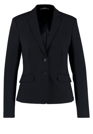 Frau trägt einen schwarzen Blazer und Rock mit weißem Spitzentop, schwarze transparente Strumpfhosen und schwarze High Heels, hält eine braune Clutch beim Gehen.