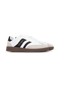 Sneakers bianchi con strisce di accento nere e dorate, dotate di un cappuccio in suede e una suola in gomma marrone. I lacci sono bianchi, realizzati con materiali testurizzati.